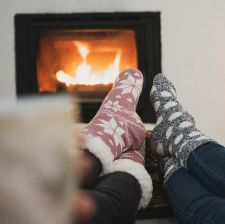 pogled s leđa dviju djevojaka koje sjede kraj kamina i božićnog drvca s nogama sa zimskim čarapama na stoliću, pijući kavu ili čaj